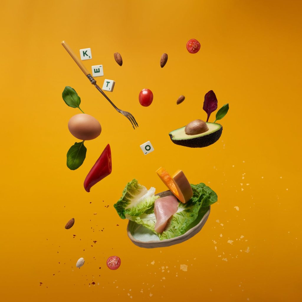 Verschillende groenten en fruit zoals tomaten en avocado's, zweven in de lucht met losse letters die samen 'keto' vormen